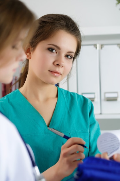 Foto duas lindas médicas trabalhando em seu escritório discutindo diagnósticos e preenchendo o registro do paciente ou formulário de histórico médico