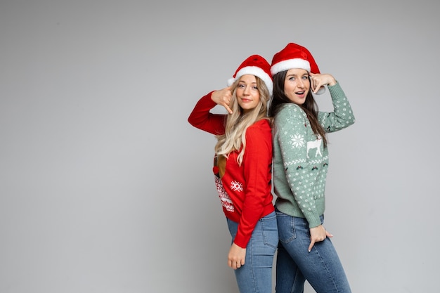 Duas lindas meninas com chapéu de Papai Noel mostram gesto de me ligar em fundo cinza com espaço de cópia