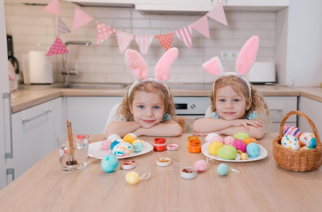 Duas lindas meninas gêmeas estão sentadas em uma mesa festiva enquanto se preparam para o feriado da Páscoa assistindo a câmera se fechar