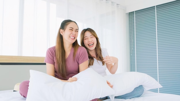 Duas lindas mulheres asiáticas em conversas casuais sorrindo e rindo com diversão enquanto estão sentadas com um travesseiro branco em uma cama em um quarto aconchegante Amizade amiga