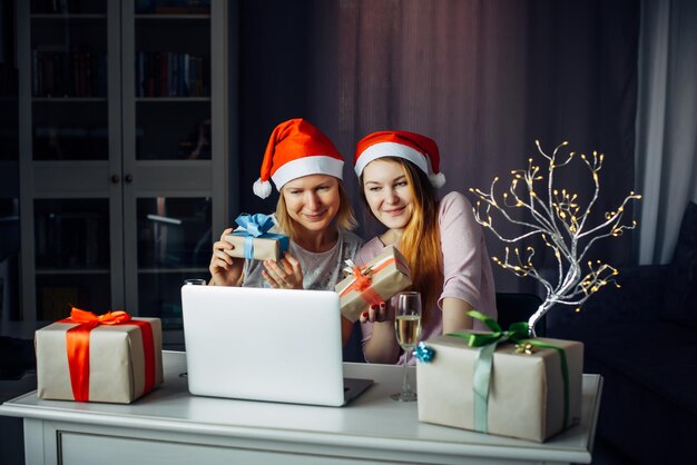 Duas lindas mulheres com chapéus de Papai Noel, sentado à mesa com o laptop entre guirlandas e presentes e desejam um feliz Natal. Comunicação com amigos e familiares via Internet na Réveillon de 2022.