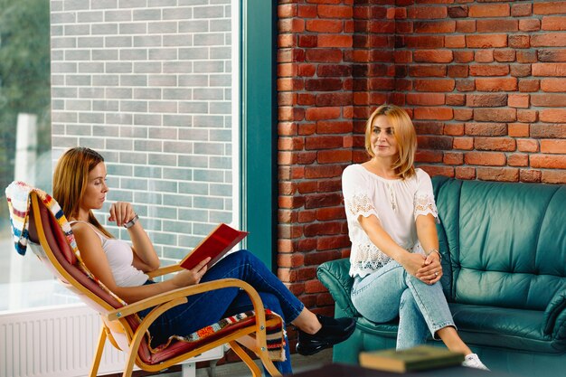 Duas lindas mulheres em um ambiente aconchegante com um livro, em uma cadeira de balanço e conversando com um bom amigo. belo interior com grandes janelas.