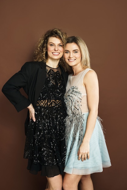 Duas lindas mulheres sorridentes com vestidos de festa brilhantes sobre fundo marrom