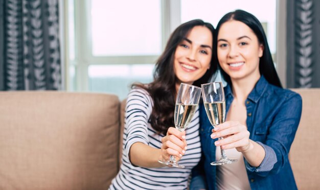 Duas lindas namoradas em roupas casuais descansam sentadas no sofá conversando e bebendo champanhe