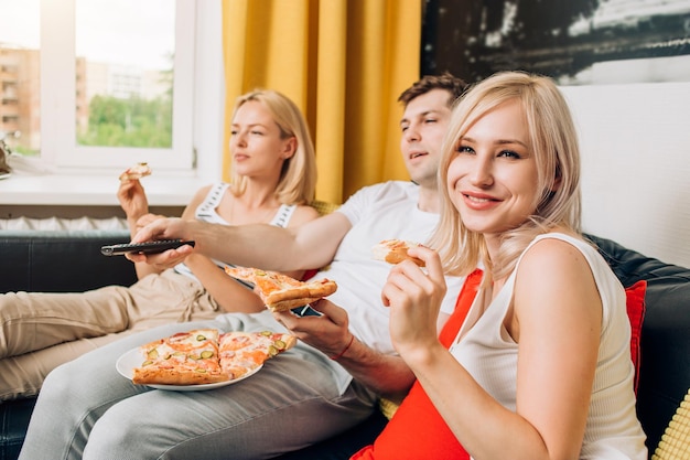 Duas loira linda mulher caucasiana e homem relaxando em casa, assistindo tv, sentado no sofá e saboreando uma pizza saborosa. foco seletivo. fast food, prazer, conceito de relaxamento.