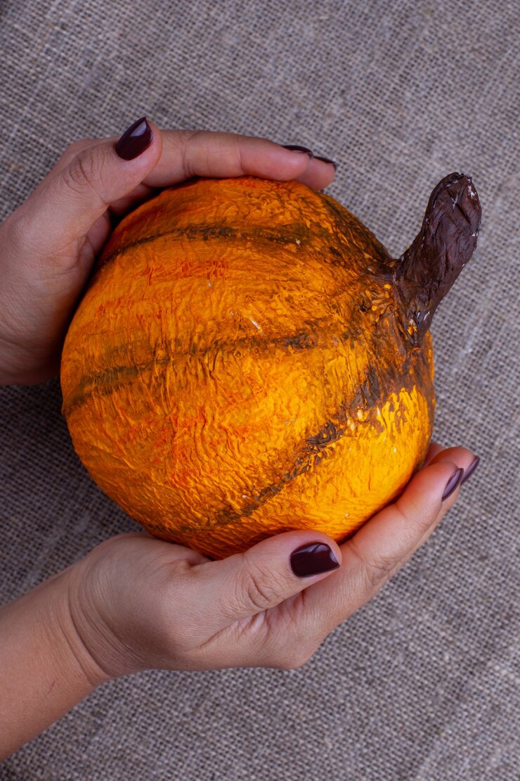 Foto duas mãos com uma manicure marrom segurando uma abóbora de papel machê laranja caseira para o halloween em uma superfície de estopa, horizontal
