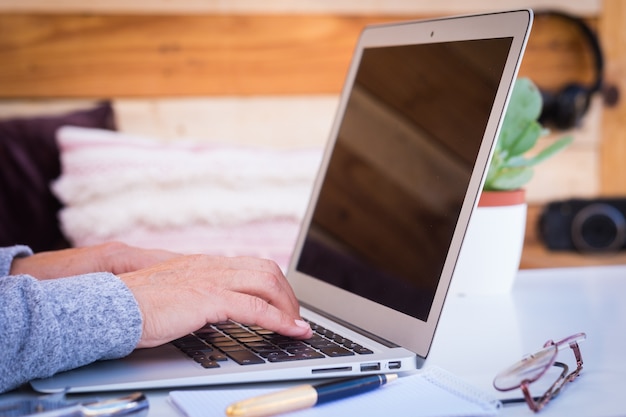 Duas mãos de uma mulher idosa trabalhando em um laptop ao ar livre em uma mesa branca com dispositivos e câmera