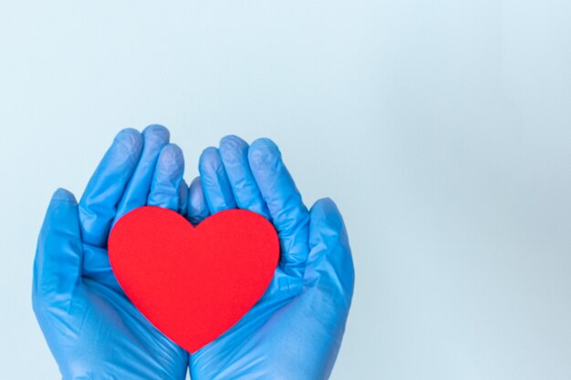 Duas mãos em luvas médicas azuis segurando uma forma de coração vermelho sobre um fundo azul, copie o espaço. Amor, conceito de saúde.