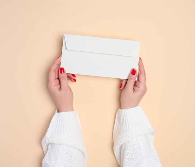 Duas mãos femininas segurando um envelope retangular branco em uma vista superior de fundo bege