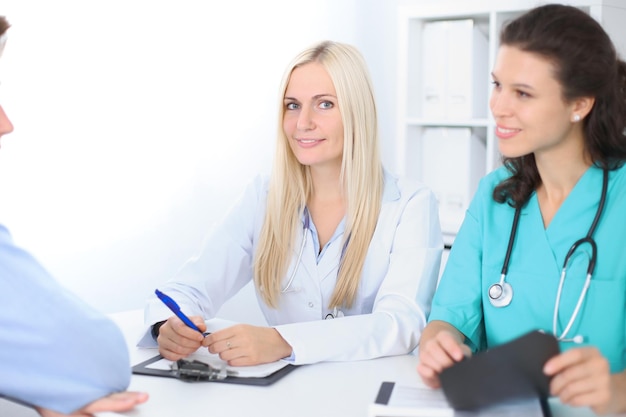 Duas médicas amigáveis confiantes sentadas à mesa e ouvem o paciente. Concentre-se na médica loira. Conceito médico e de saúde.