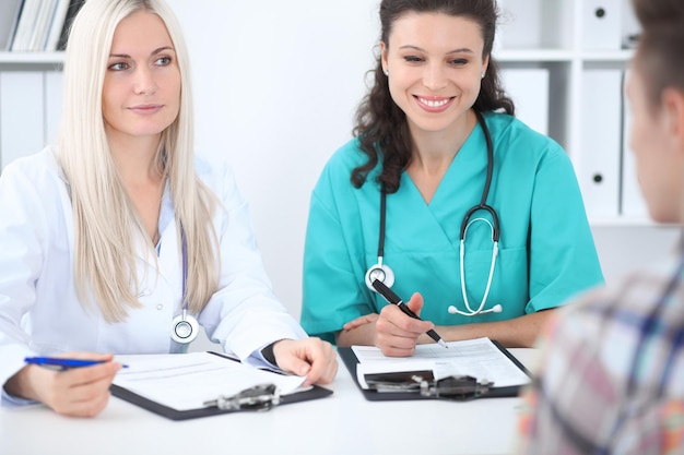 Duas médicas amigáveis confiantes sentadas à mesa e ouvem o paciente. Concentre-se na médica loira. Conceito médico e de saúde.