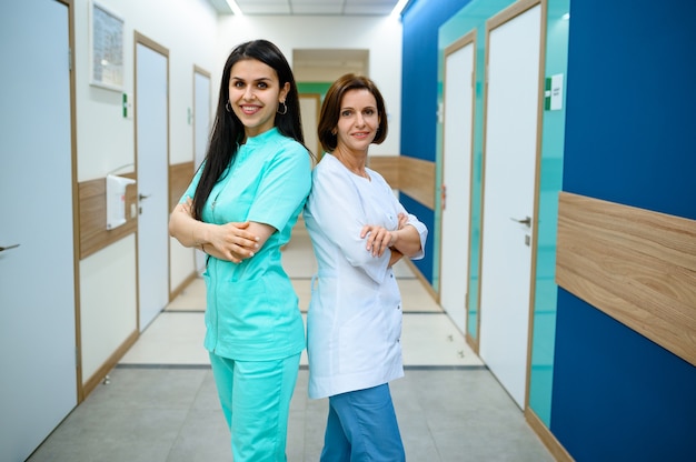 Duas médicas sorridentes em poses uniformes no corredor da clínica. Profissional médico especialista em hospital, laringologista ou otorrinolaringologista, ginecologista ou mamologista, cirurgião