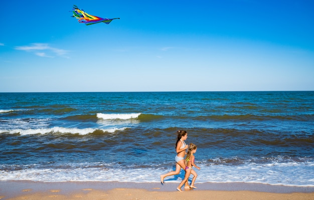 Duas meninas alegres felizes correm com uma pipa na costa arenosa à beira-mar. Dia quente de verão ensolarado. Conceito de jogos infantis ativos. Copyspace