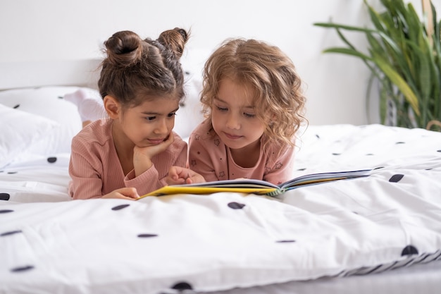 Duas meninas amigas de pijama lendo um livro deitado em uma cama branca na cama em casa