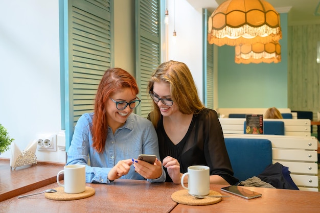 Duas meninas animadas usando telefones celulares sentadas em um café e apontando o dedo Mulher ruiva em óculos mostra a sua amiga fotos engraçadas em um smartphone namorada loira Eles bebem café