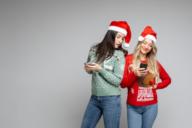 Duas meninas com gorros de Papai Noel e suéteres de inverno fazem pedidos, compram on-line por telefone em fundo cinza, copie o espaço