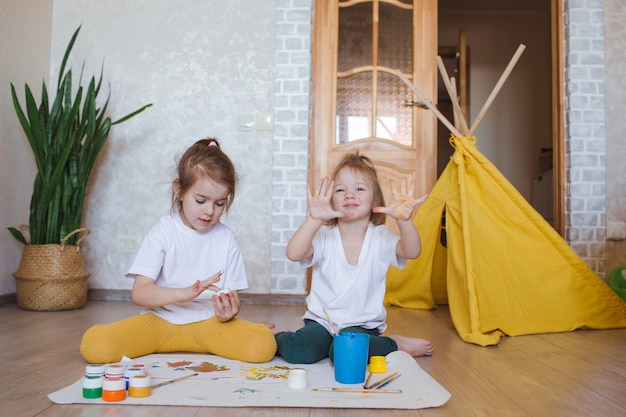 Duas meninas com roupas de cores claras sentam-se no chão de joelhos e desenham com entusiasmo aquarelas brilhantes.