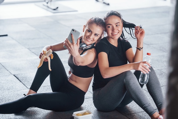 Foto duas meninas com roupas esportivas, sentadas com comida na academia juntos durante o dia e fazendo uma selfie.