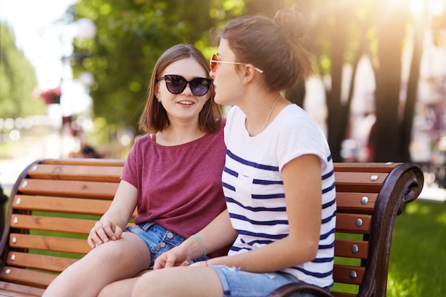 Duas meninas criativas falam e riem enquanto está sentado no banco ao ar livre.