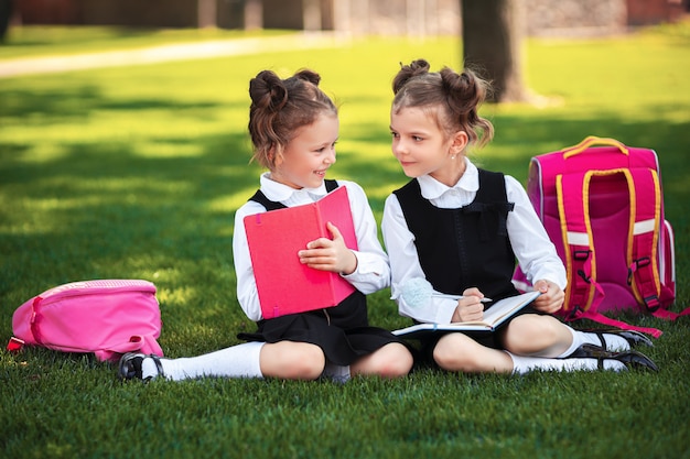 Duas meninas da escola com mochila rosa sentada na grama depois das aulas e ler livro ou estudar lições,