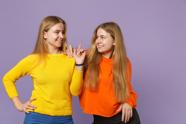 Duas meninas de irmãs gêmeas jovens loiras em roupas coloridas, olhando uma para a outra, apontando o dedo indicador isolado na parede azul violeta. Conceito de estilo de vida familiar de pessoas.