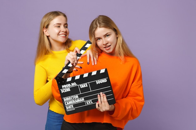 Duas meninas de irmãs gêmeas loiras jovens em roupas coloridas, segurando o clássico filme preto, fazendo claquete isolada na parede azul violeta. Conceito de estilo de vida familiar de pessoas. .