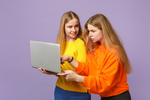 Duas meninas de irmãs gêmeas loiras jovens encantadoras em roupas coloridas vivas segurando usando computador laptop pc isolado na parede azul violeta pastel. Conceito de estilo de vida familiar de pessoas.