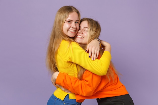 Duas meninas de irmãs gêmeas loiras muito alegres em roupas coloridas vivas, abraçando-se isoladas na parede de azul violeta pastel. conceito de estilo de vida familiar de pessoas.