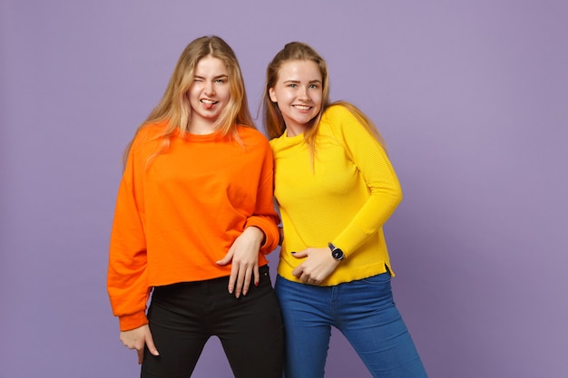 Duas meninas de irmãs gêmeas muito jovens loiras em roupas coloridas vivas, mostrando a língua isolada na parede de azul violeta pastel. conceito de estilo de vida familiar de pessoas.