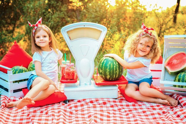 Duas meninas em uma camiseta branca e uma bandana vermelha sentam-se em um tapete na natureza e seguram uma melancia e uma jarra com suco, que fica em uma balança à moda antiga. conceito de vendas