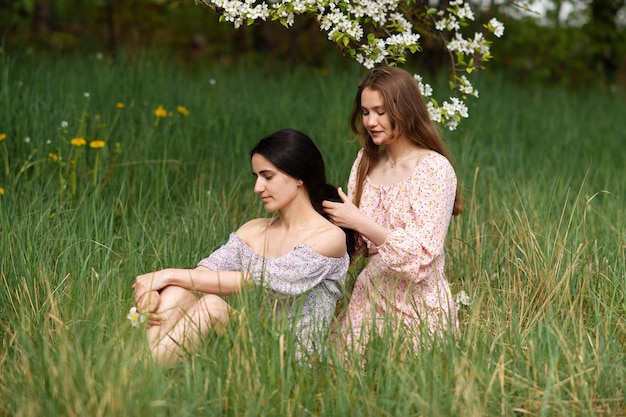 Duas meninas em vestidos estão sentadas debaixo de uma árvore branca e trançando o cabelo uma da outra