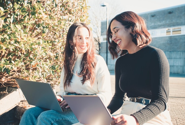 Duas meninas estudando em laptops enquanto conversam e riem ao ar livre da faculdade durante um dia brilhante. Estudos universitários, trabalhos universitários, estilo moderno. Preparando uma apresentação para a aula.