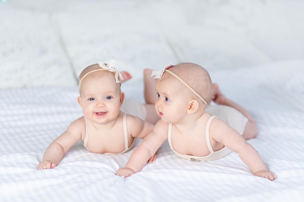 Duas meninas gêmeas recém-nascidas em um terno de algodão em uma cama branca em casa deitam-se de bruços e sorriem enquanto brincam uma com a outra