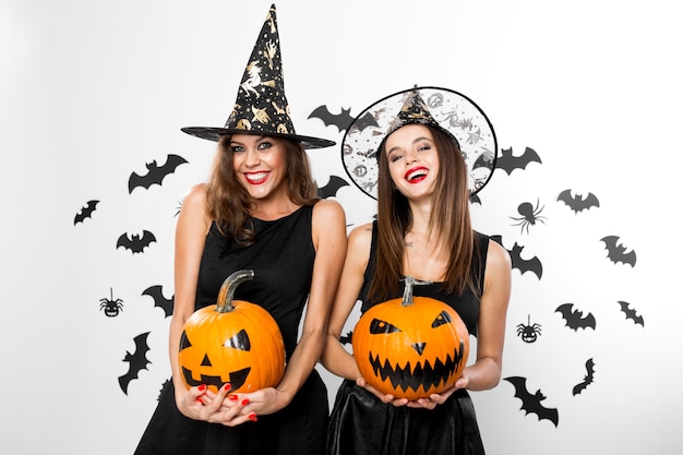 Duas meninas morenas em vestidos pretos e chapéus de bruxa seguram abóboras de halloween de terror no fundo com morcegos.