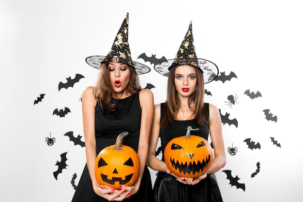 Duas meninas morenas em vestidos pretos e chapéus de bruxa seguram abóboras de halloween de terror no fundo com morcegos.
