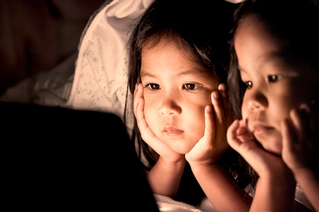 Duas meninas pequenas asiáticas usando o tablet digital sob o cobertor à noite no tom de cor vintage