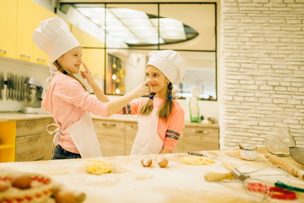 Duas meninas sorridentes cozinheiros de bonés e aventais, se divertindo, preparação de biscoitos na cozinha. crianças cozinhando massa, crianças chefs preparando bolo