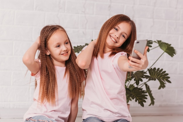 Duas meninas sorriem alegremente e tiram uma foto de selfie em um smartphone em uma sala branca