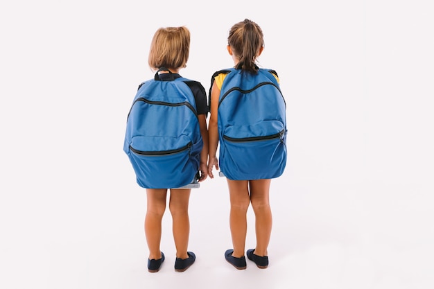 Duas meninas, uma de cabelos louros e a outra de cabelos pretos, vestidas com macacão jeans azul, com mochila, prontas para a volta às aulas, de costas voltadas, sobre fundo branco.