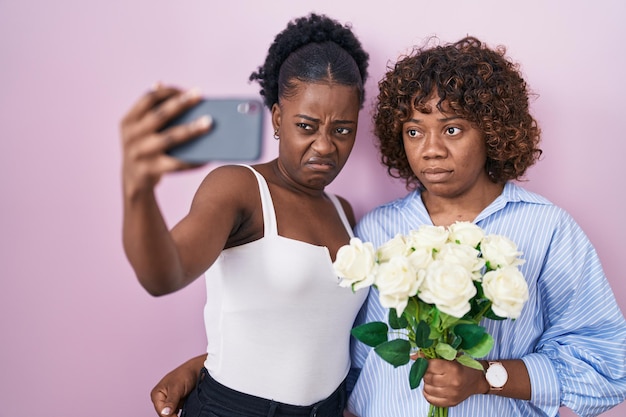 Duas mulheres africanas tirando uma foto selfie com flores céticas e nervosas, franzindo a testa chateadas por causa do problema. pessoa negativa.