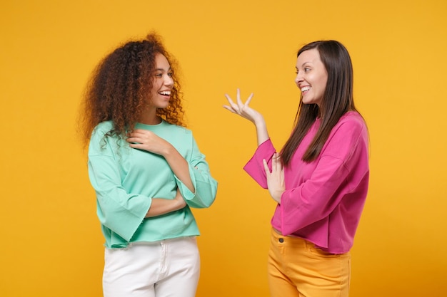 Foto duas mulheres alegres amigas europeias e afro-americanas em roupas verdes rosa posando isoladas em fundo amarelo. conceito de estilo de vida de pessoas. mock up espaço de cópia. olhando um para o outro falando.