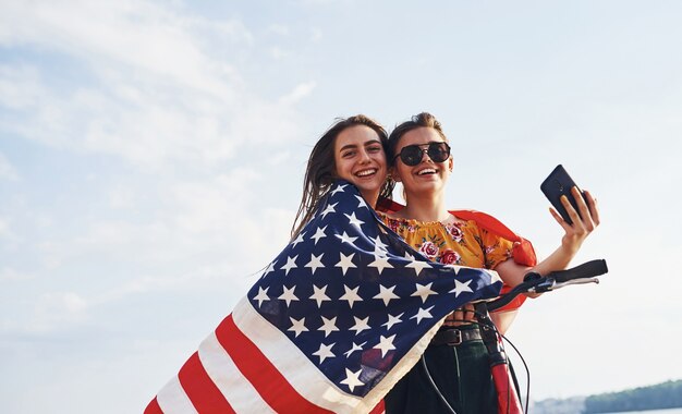 Duas mulheres alegres patrióticas com bicicleta e a bandeira dos EUA nas mãos fazem selfie.