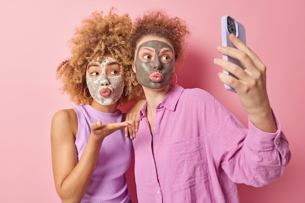 Duas mulheres aplicam máscaras de beleza no rosto sopro beijo de ar na câmera frontal do smartphone passam por procedimentos de cuidados com a pele vestidos com roupa casual isolada sobre fundo rosa Conceito de tratamentos faciais