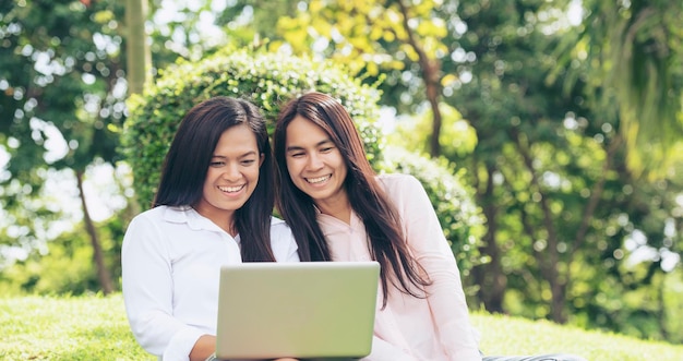 Duas mulheres asiáticas conversando usando laptop sentado no prado verde no parque amizade meninas conversam e conversam olham para laptop planejando fazer pequenos negócios jardim verde jovens mulheres bonitas riem conversam juntas