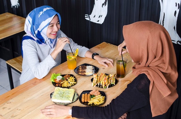 Duas mulheres asiáticas hijab comendo sua comida e rindo com seus amigos em um restaurante lotado