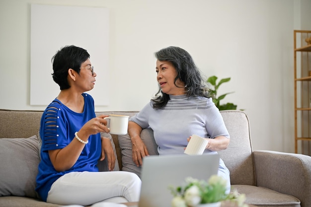 Duas mulheres asiáticas maduras de meia-idade estão desfrutando de seu café da manhã e conversando em um sofá
