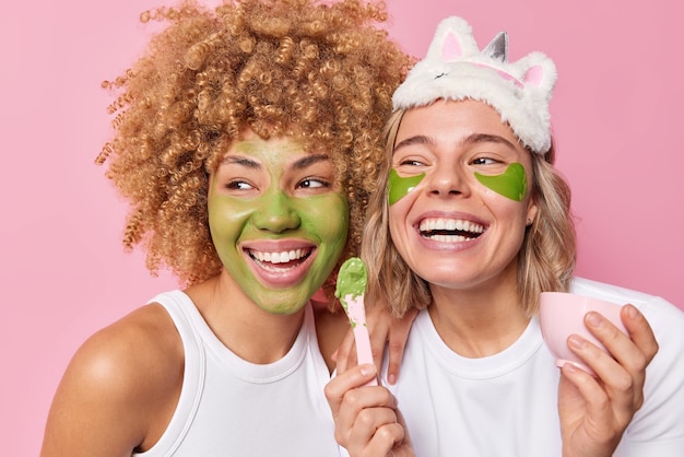 Duas mulheres atraentes passam por procedimentos de beleza aplicam máscara verde e patches cuidam do sorriso da pele amplamente vestidos com roupas casuais ficam um ao lado do outro isolado sobre fundo rosa