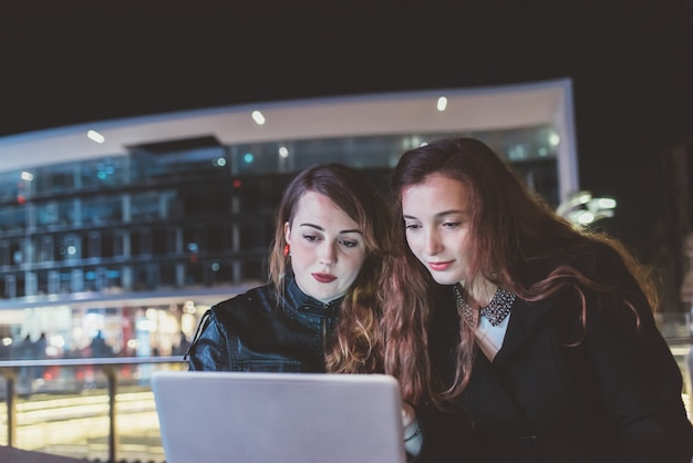 Duas mulheres bonitas ao ar livre à noite, usando o computador