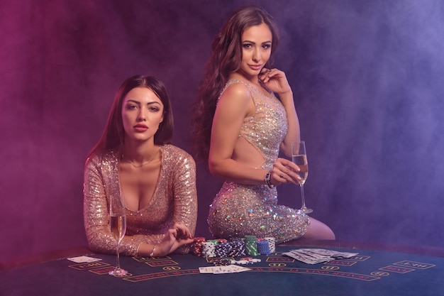 Duas mulheres bonitas em vestidos dourados bebendo champanhe posando na mesa de jogo no cassino Luzes de fundo coloridas de fundo de fumaça preta Jogo de entretenimento fichas de pôquer cartões de dinheiro Closeup