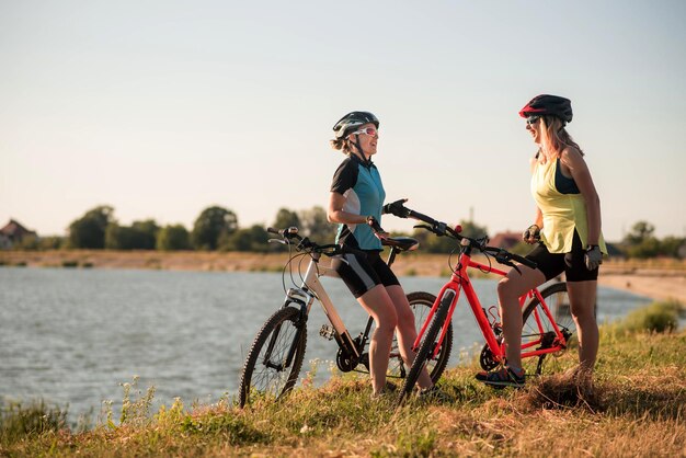 Duas mulheres ciclistas com bicicletas em pé e conversando na margem do lago